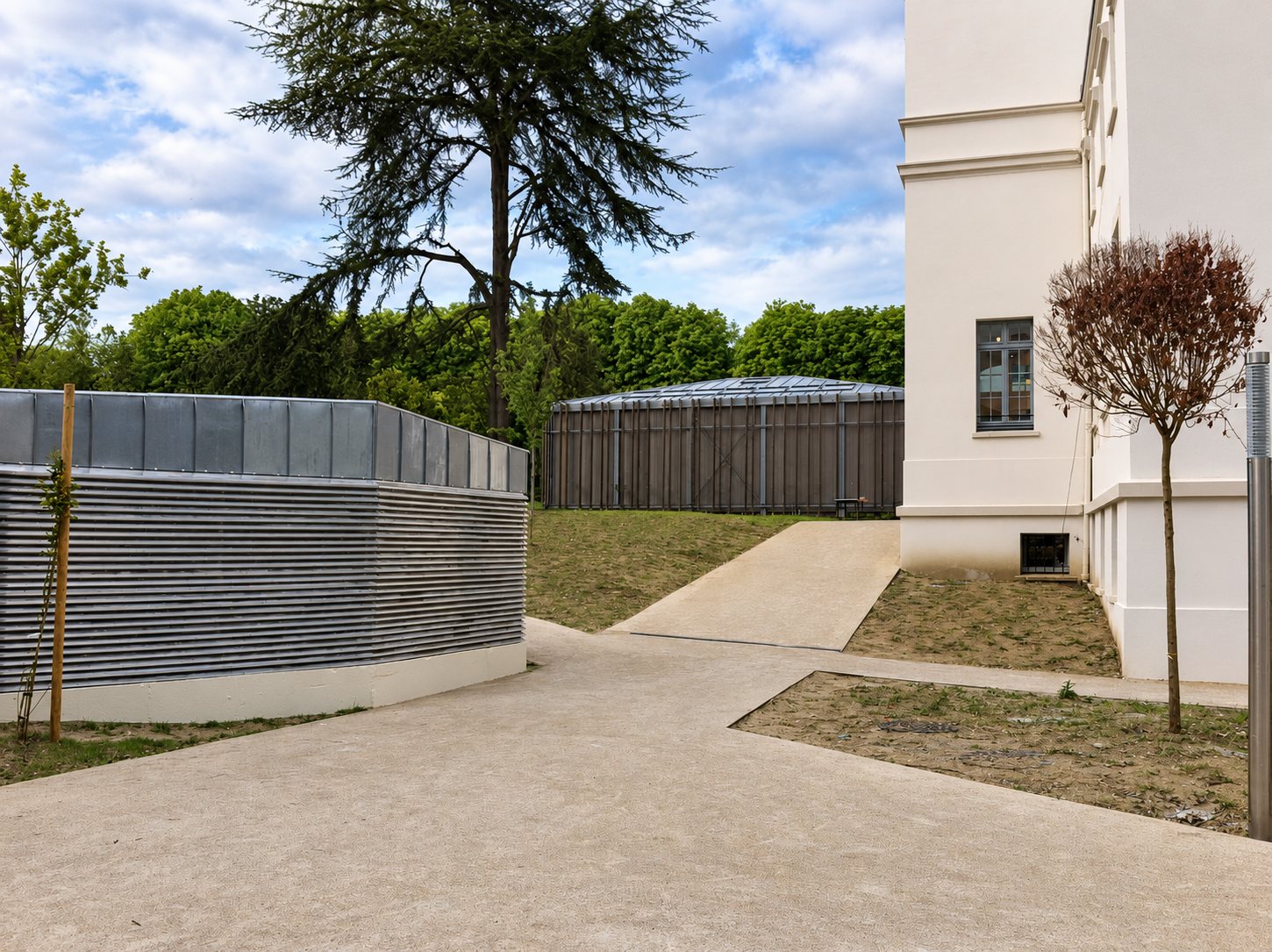 Cour fraîchement aménagée avec des allées en béton, de jeunes arbres et un local à parois métalliques, à côté d’un bâtiment blanc et d’un grand cèdre en arrière-plan.