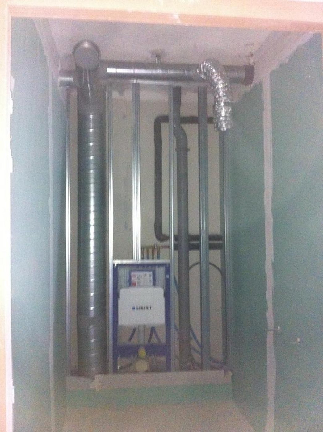 Un bâti-support Geberit pour WC suspendu est monté entre des rails métalliques, avec des conduits de ventilation et des tuyaux d’évacuation, dans une gaine entourée de plaques de plâtre vertes.