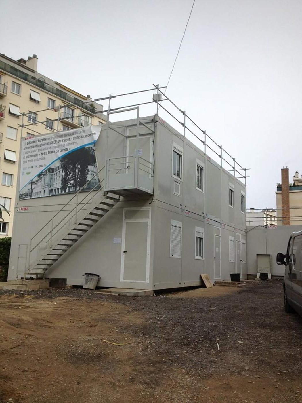 Un bâtiment modulaire gris sur deux niveaux, équipé d’un escalier métallique et d’une grande bâche publicitaire, installé sur un terrain de chantier entre des immeubles.