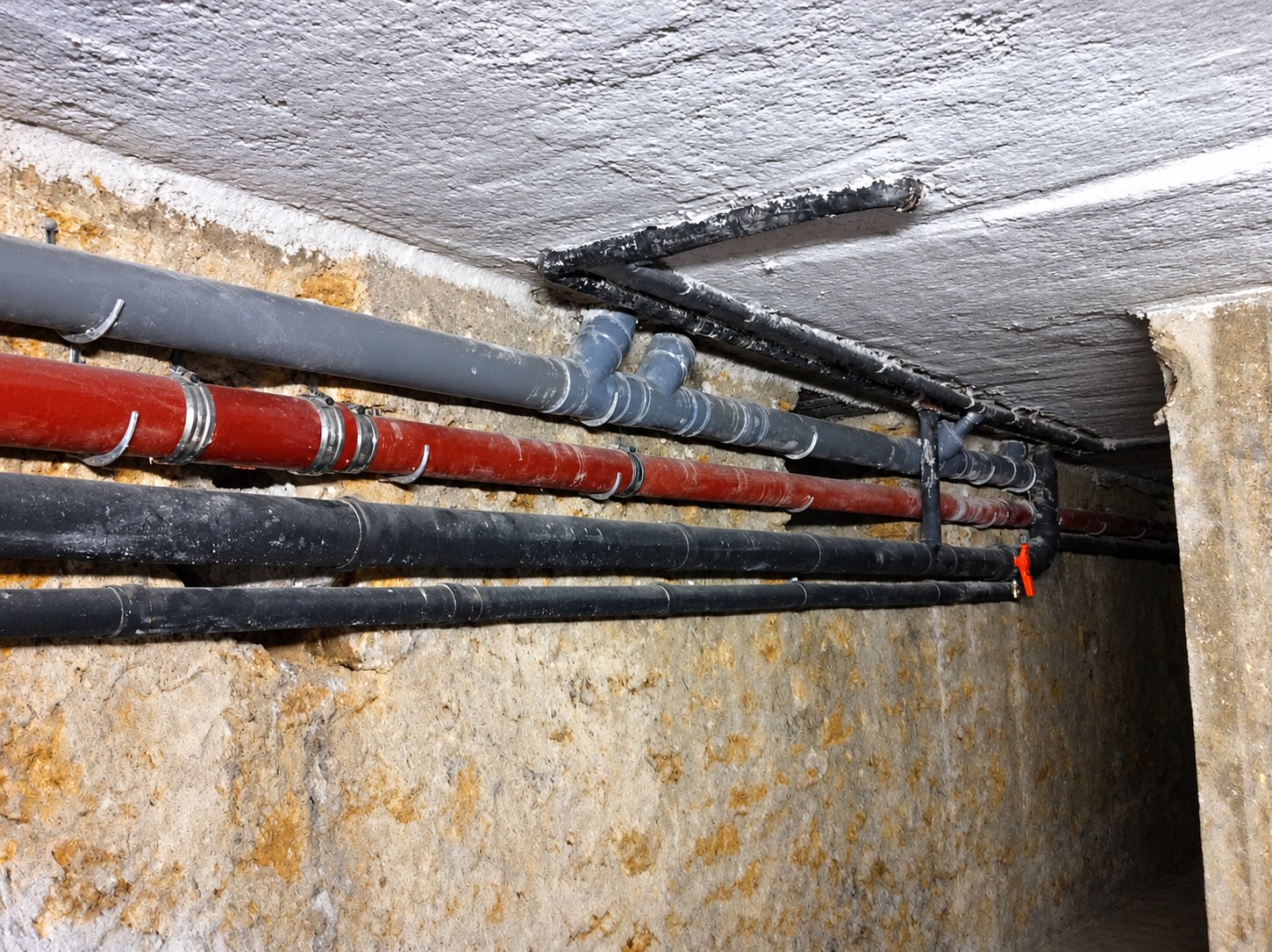 Plusieurs tuyaux de plomberie gris, rouge et noirs fixés par des colliers sous un plafond en béton, le long d’un mur de pierre dans une cave.