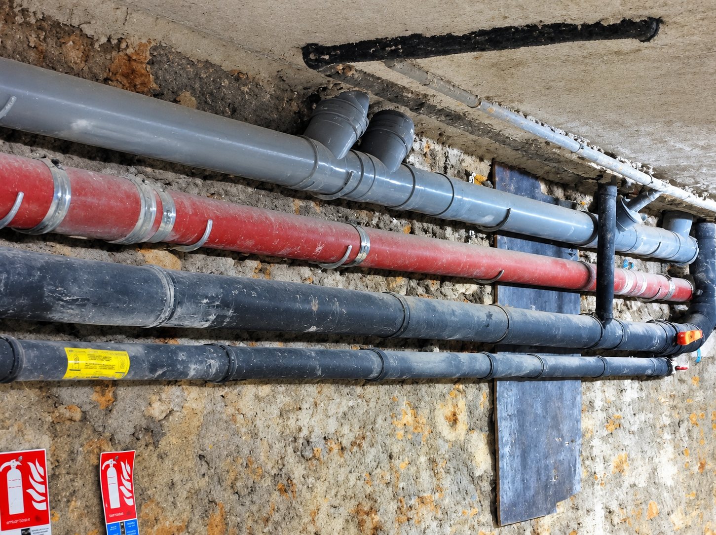 Plusieurs tuyaux gris, rouge et noirs courent le long d’un plafond en béton, avec deux pictogrammes d’extincteur rouges fixés au mur.