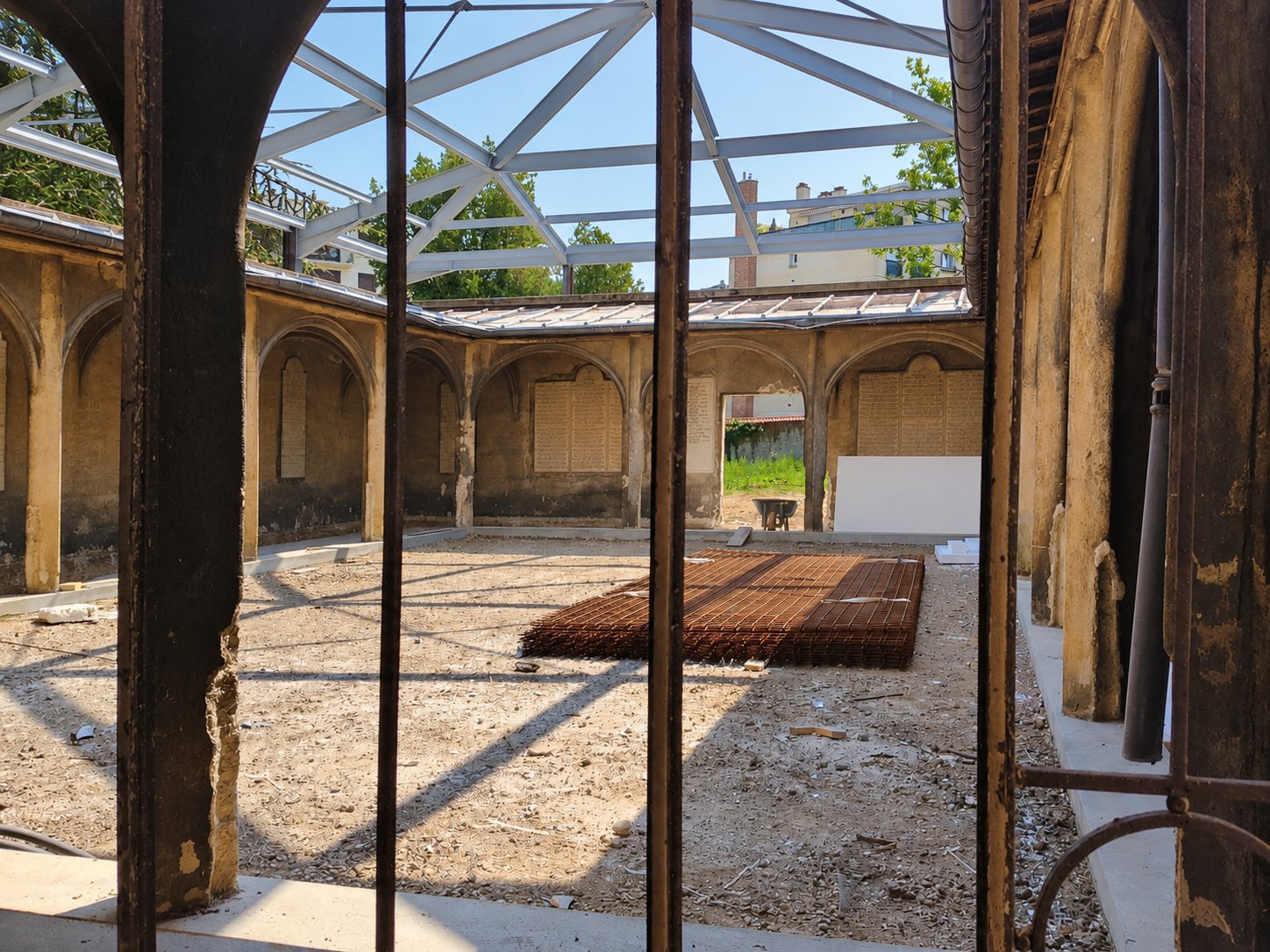 Vue à travers une grille d’une cour à arcades en rénovation, avec une structure métallique en toiture et un treillis d’armatures posé sur le sol de terre.