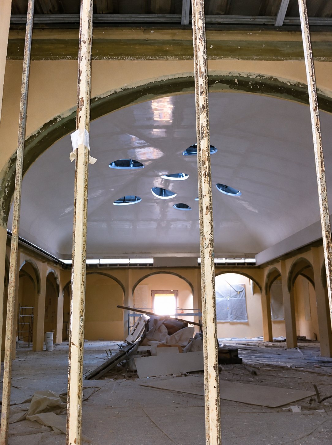 Vue à travers des barreaux sur une grande salle voûtée en travaux, avec un plafond tendu blanc percé d’ouvertures ovales et des gravats au sol.
