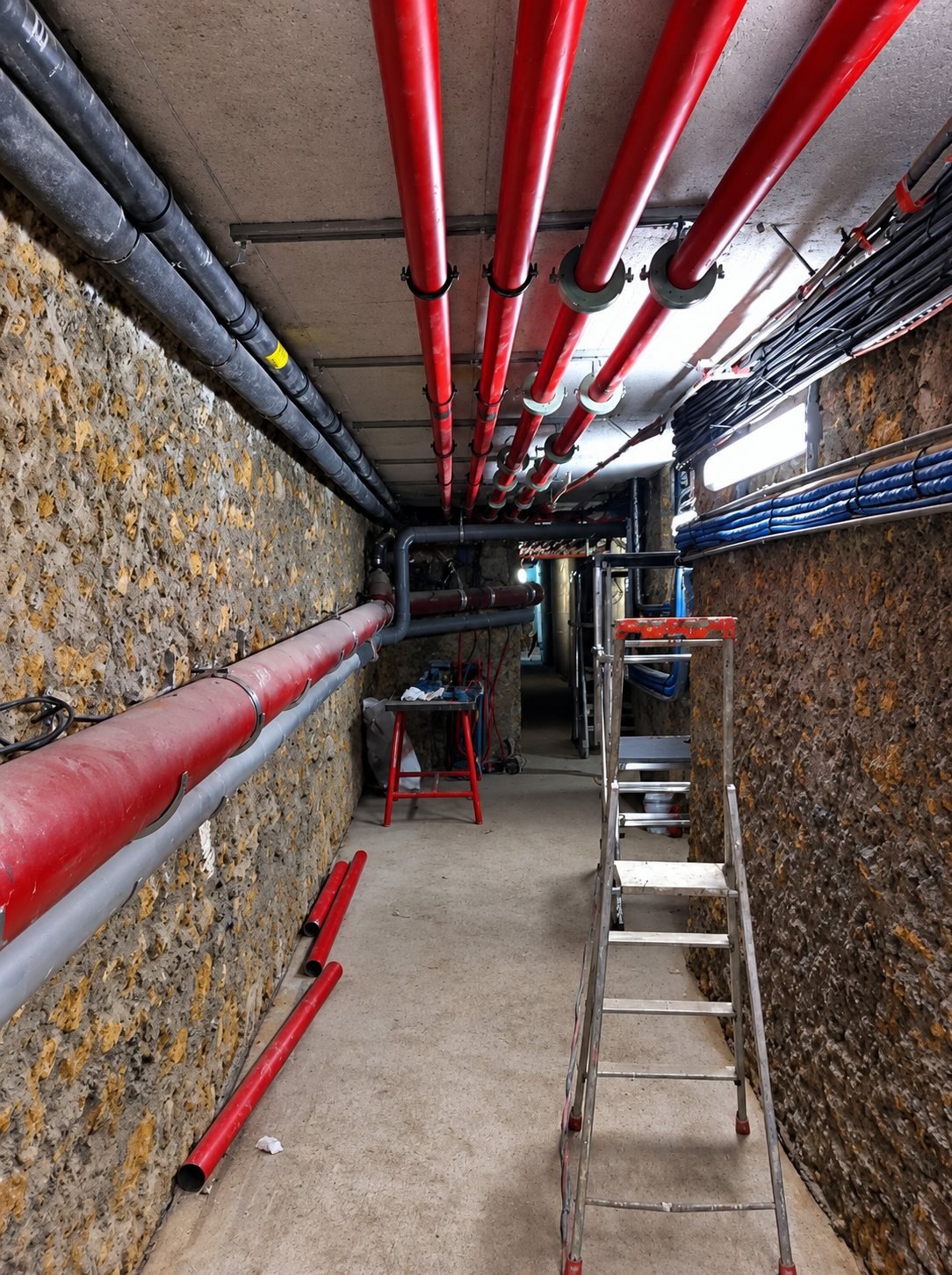 Un couloir souterrain en pierre où des tuyaux rouges et des câbles courent au plafond et le long des murs, avec une échelle et des tronçons de tuyau posés au sol.