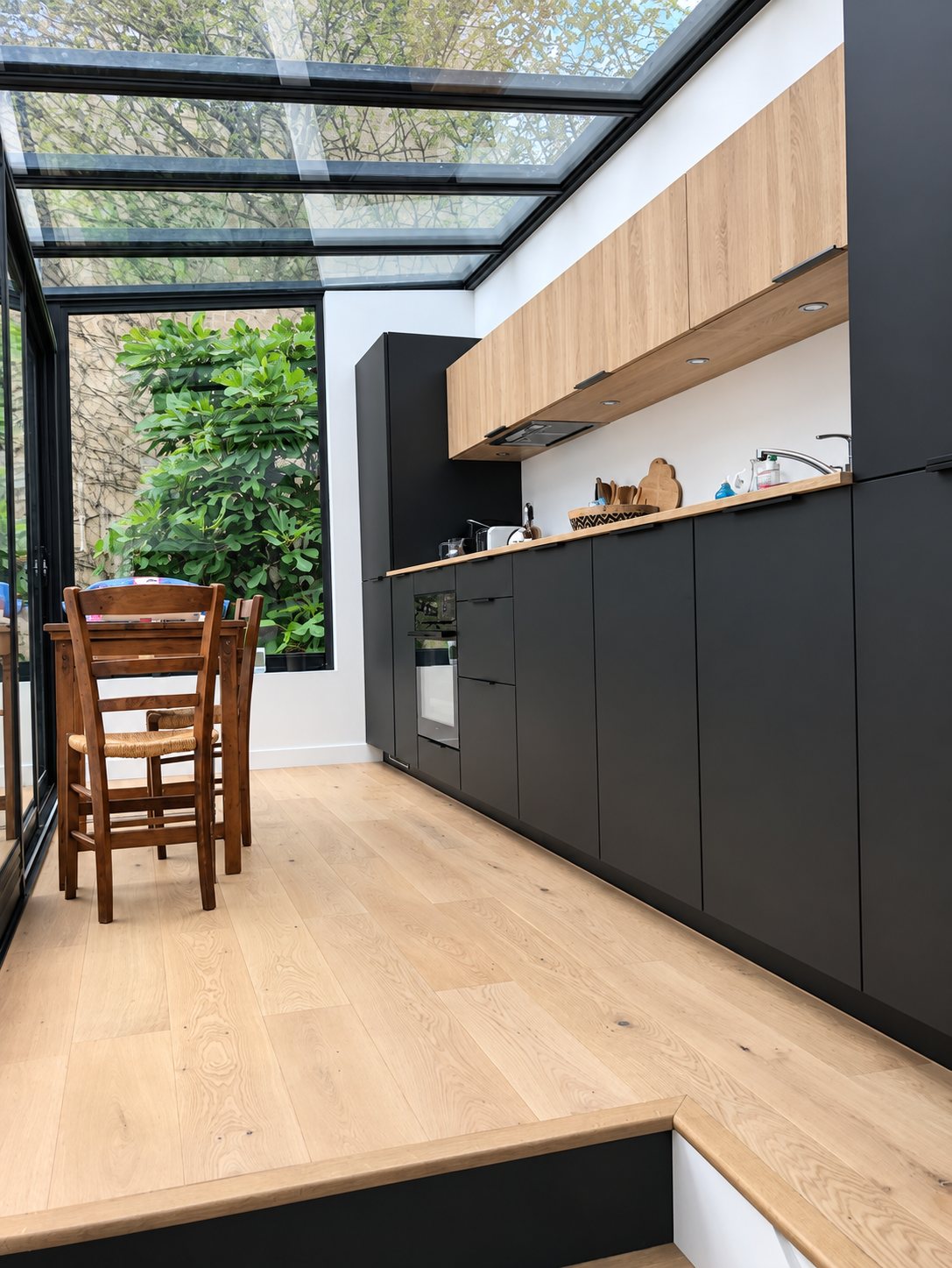 Cuisine contemporaine aux façades noires mates et meubles hauts en bois clair, installée sous une verrière avec une table et des chaises en bois donnant sur un jardin.