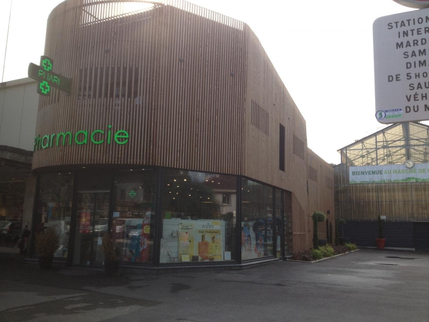La façade arrondie en lattes de bois d’une pharmacie, avec son enseigne verte, sa croix lumineuse et sa vitrine donnant sur un parking.