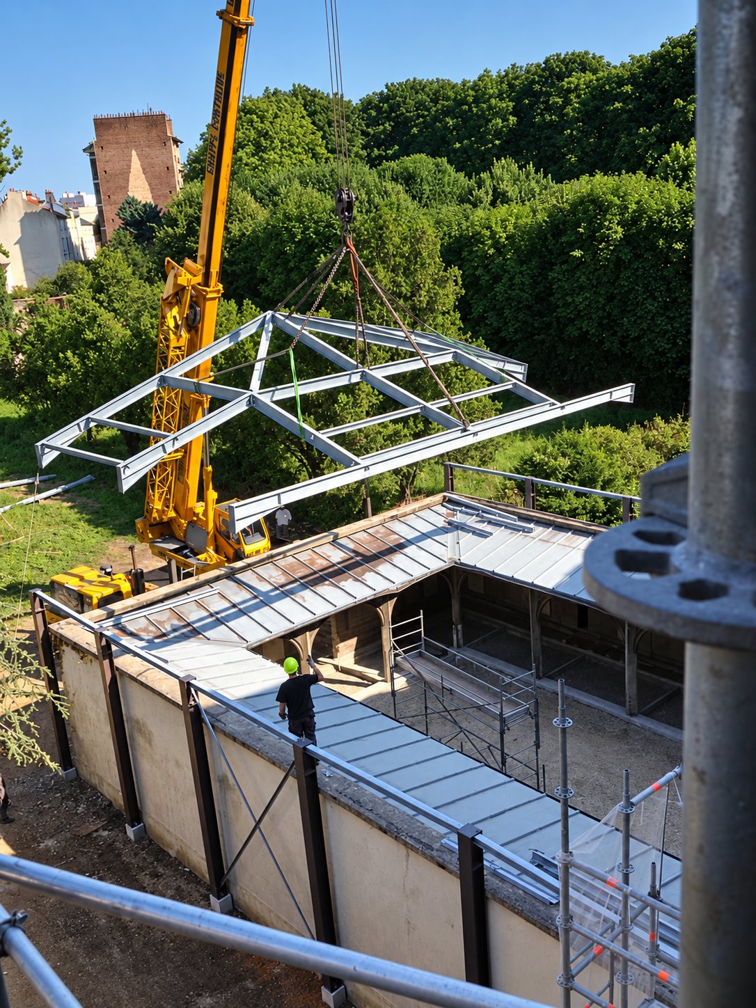 Une grue jaune soulève une charpente métallique au-dessus d’un bâtiment en chantier entouré d’échafaudages et d’arbres.