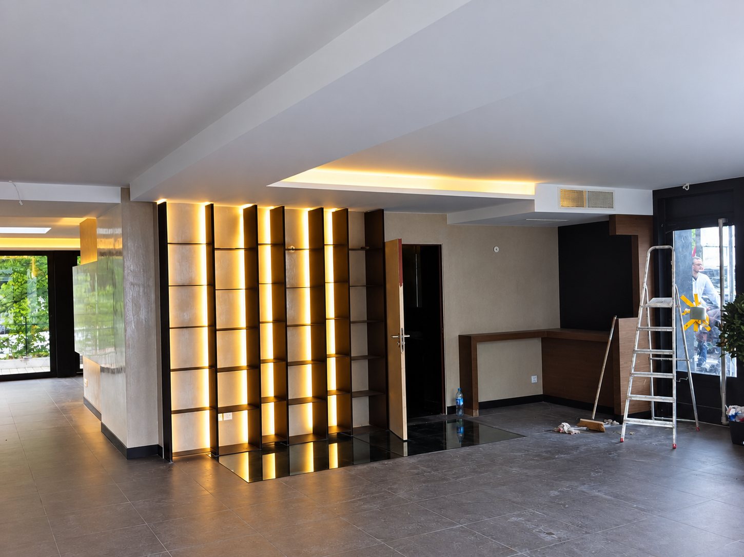 Grand hall en cours de finition avec une bibliothèque murale rétroéclairée par des LED, un plafond à éclairage indirect et une échelle posée au sol.