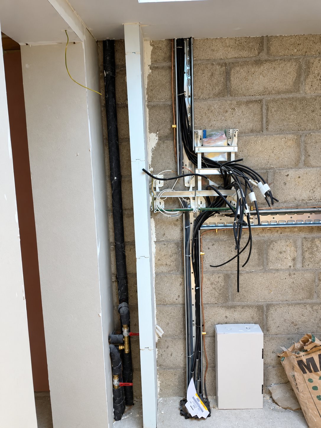 Câbles électriques noirs fixés sur des rails métalliques le long d’un mur de parpaings, à côté d’une canalisation noire et d’un petit coffret blanc sur un chantier.