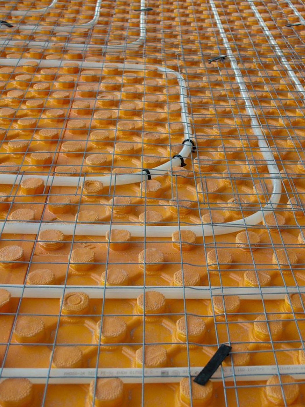 Vue rapprochée d’un chantier de plancher chauffant : tubes blancs fixés sur une plaque orange à plots et recouverts d’un treillis métallique.