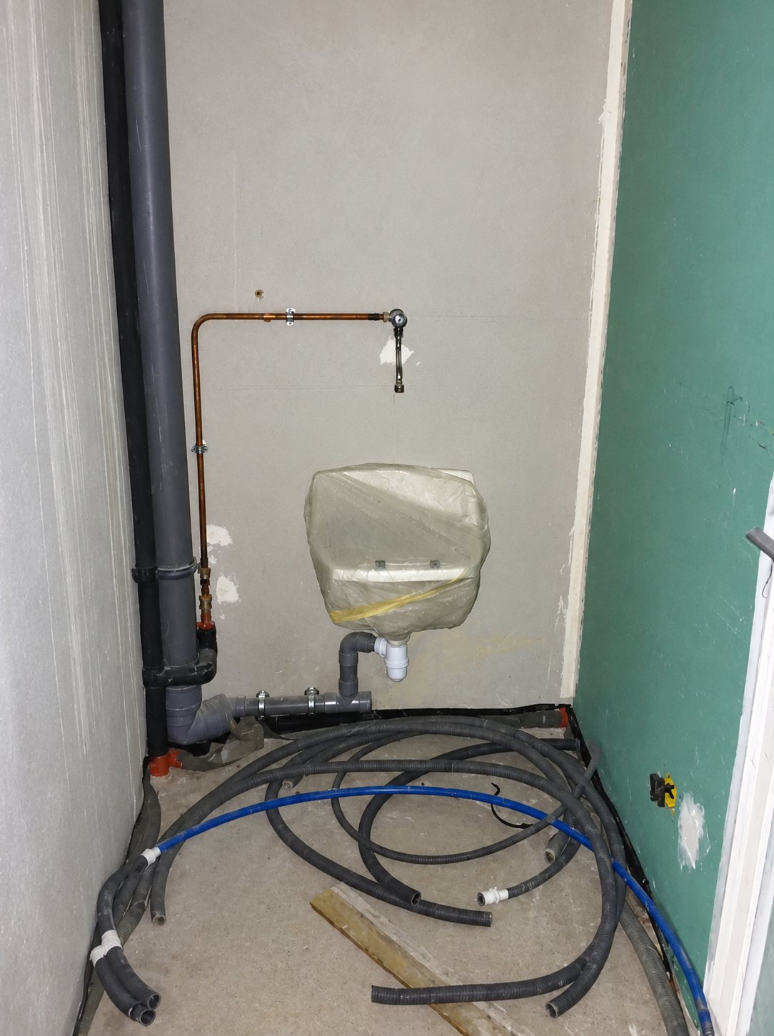 Un petit lave-mains protégé par un film plastique est fixé au mur d’un local en chantier, avec des tuyaux en cuivre, un tuyau d’évacuation gris et des gaines au sol.