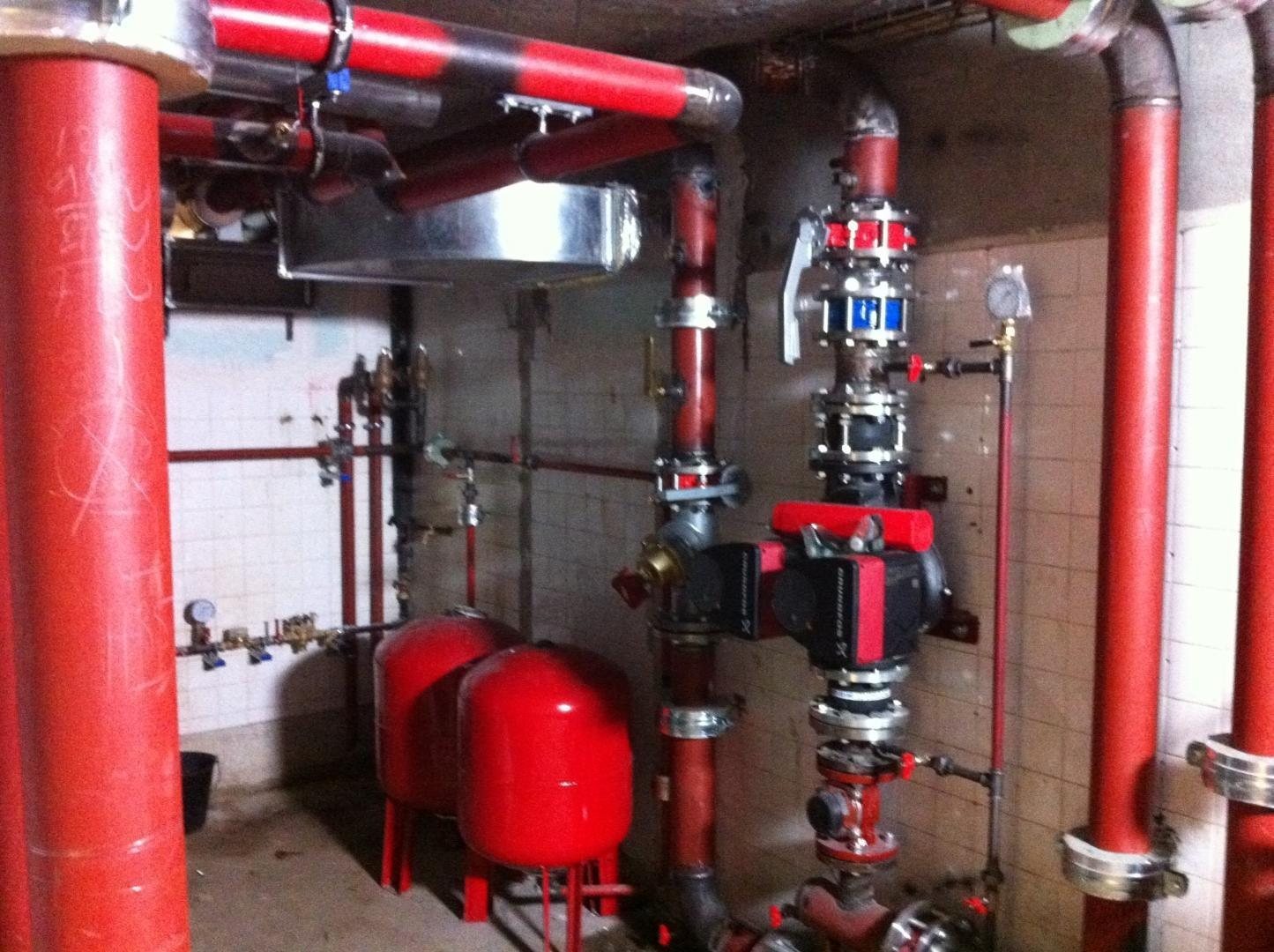 Installation de chauffage dans un local technique carrelé, avec grosses canalisations rouges, une pompe de circulation et deux vases d’expansion rouges.