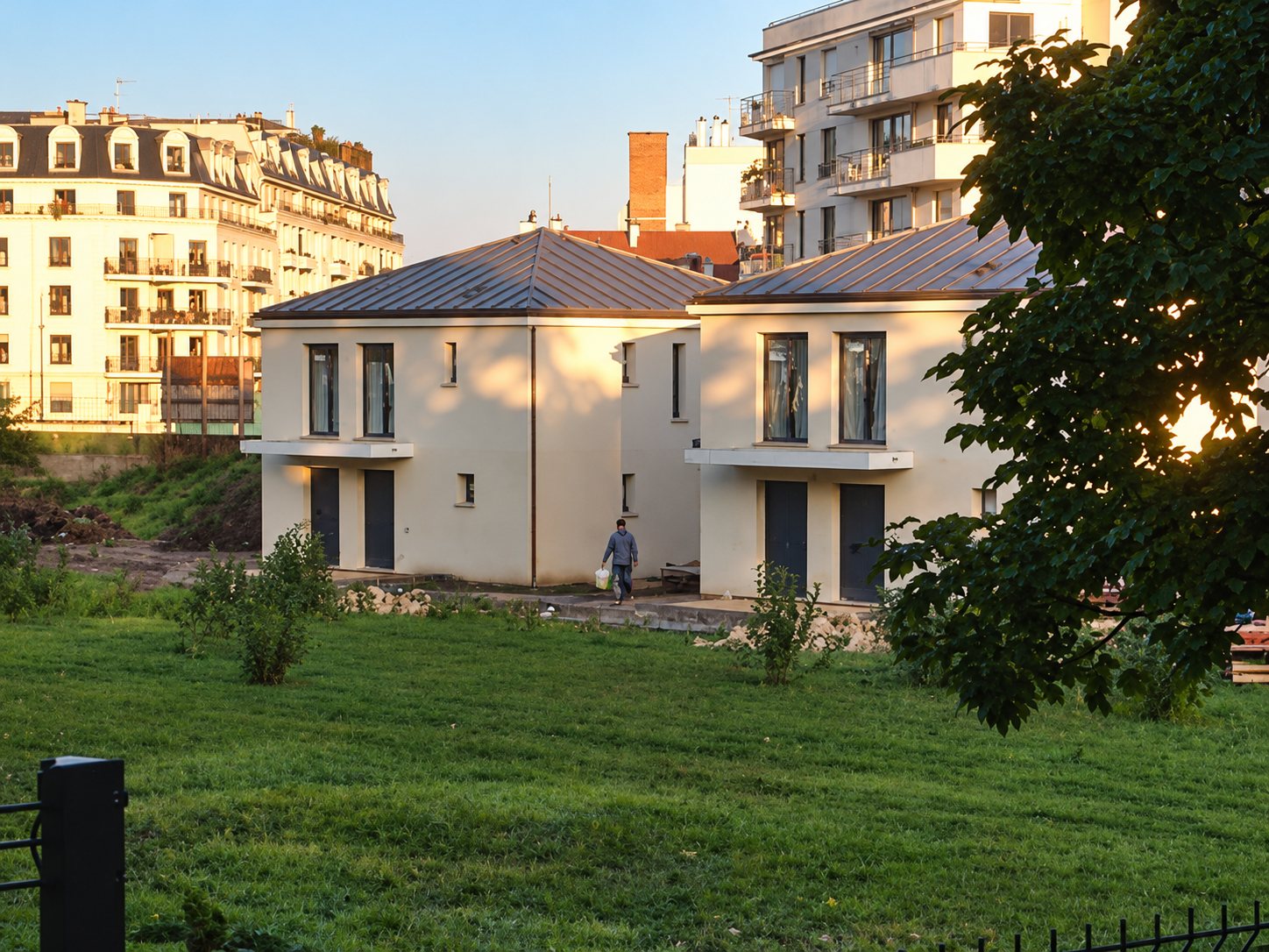 Deux maisons neuves aux façades beiges et aux toits métalliques, entourées de pelouse, avec des immeubles en arrière-plan à la lumière du soir.