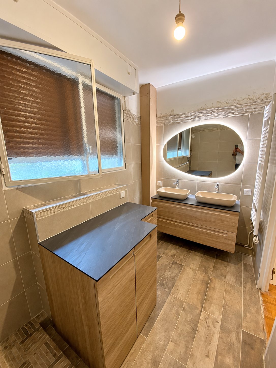 Salle de bain rénovée avec meuble double vasque en bois, miroir ovale rétroéclairé, carrelage effet bois et sèche-serviettes.