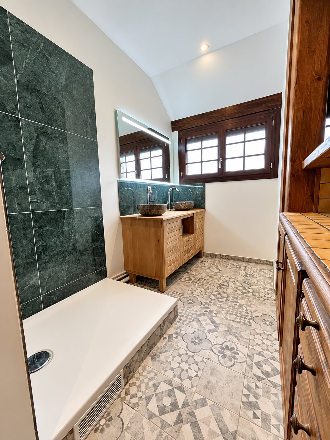 Salle de bain rénovée avec grand receveur de douche, mur en carreaux verts, meuble vasque en bois à deux vasques en pierre et sol à motifs gris.