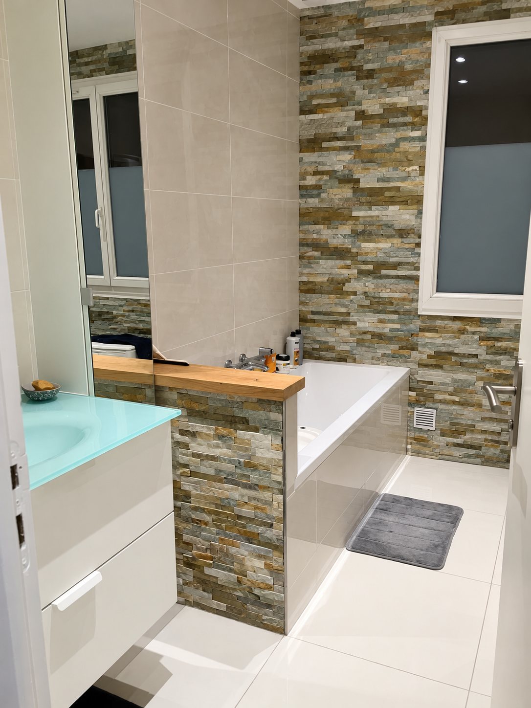 Salle de bains rénovée avec baignoire encastrée, mur en parement de pierre, vasque en verre turquoise et grand carrelage blanc au sol.