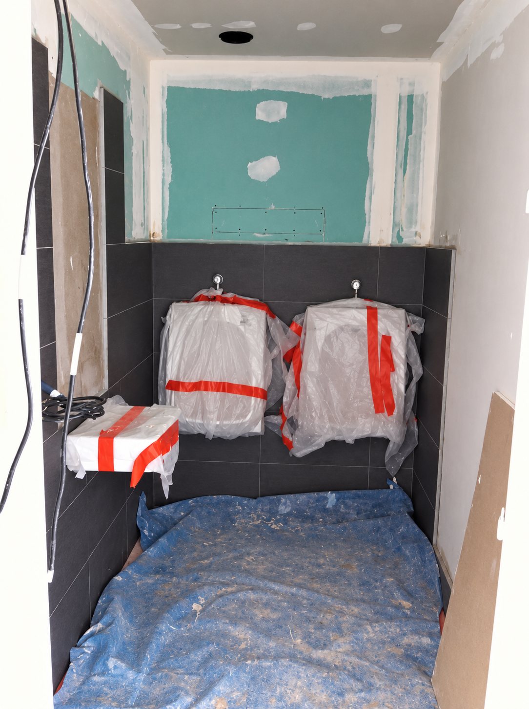Petite pièce en cours de rénovation avec carrelage gris foncé, plaques de plâtre vertes et deux sanitaires suspendus emballés dans du plastique fermé par du ruban orange.