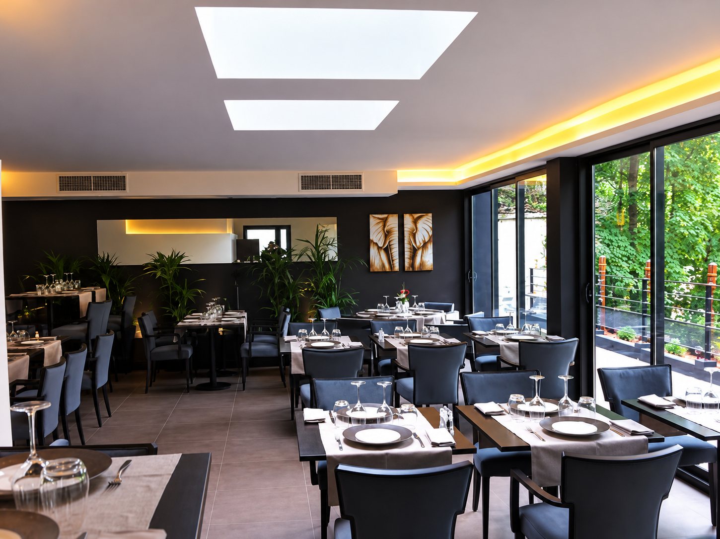 Salle de restaurant élégante aux tables dressées, chaises bleu foncé et grandes baies vitrées donnant sur un jardin verdoyant.