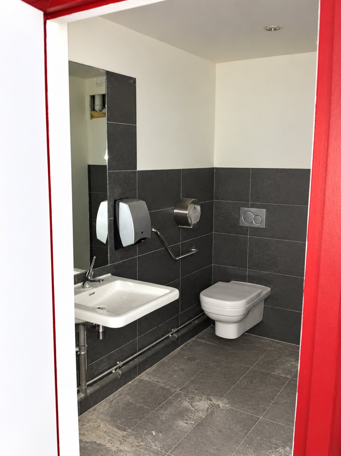 Une salle de toilettes aux murs carrelés gris foncé, équipée d’un lavabo, d’un WC suspendu et d’un distributeur d’essuie-mains, vue depuis l’encadrement rouge de la porte.