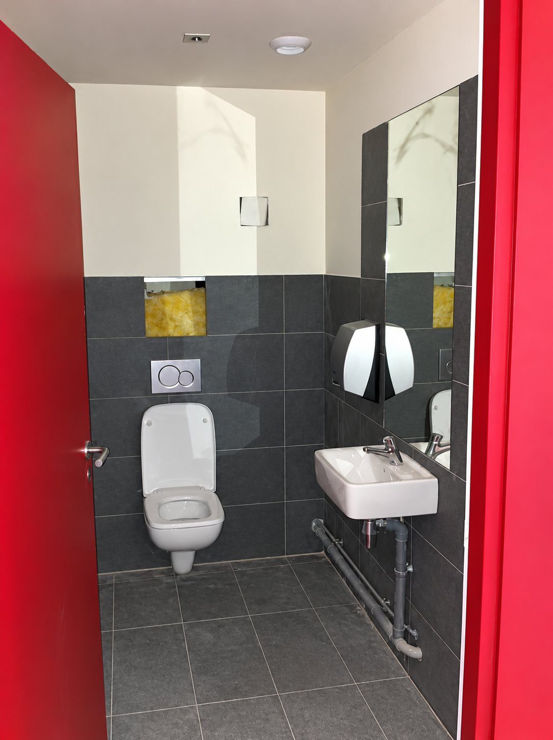 Petite salle de toilettes aux murs et au sol carrelés de gris foncé, avec un WC suspendu, un lavabo mural et un grand miroir, encadrée par des portes rouges.