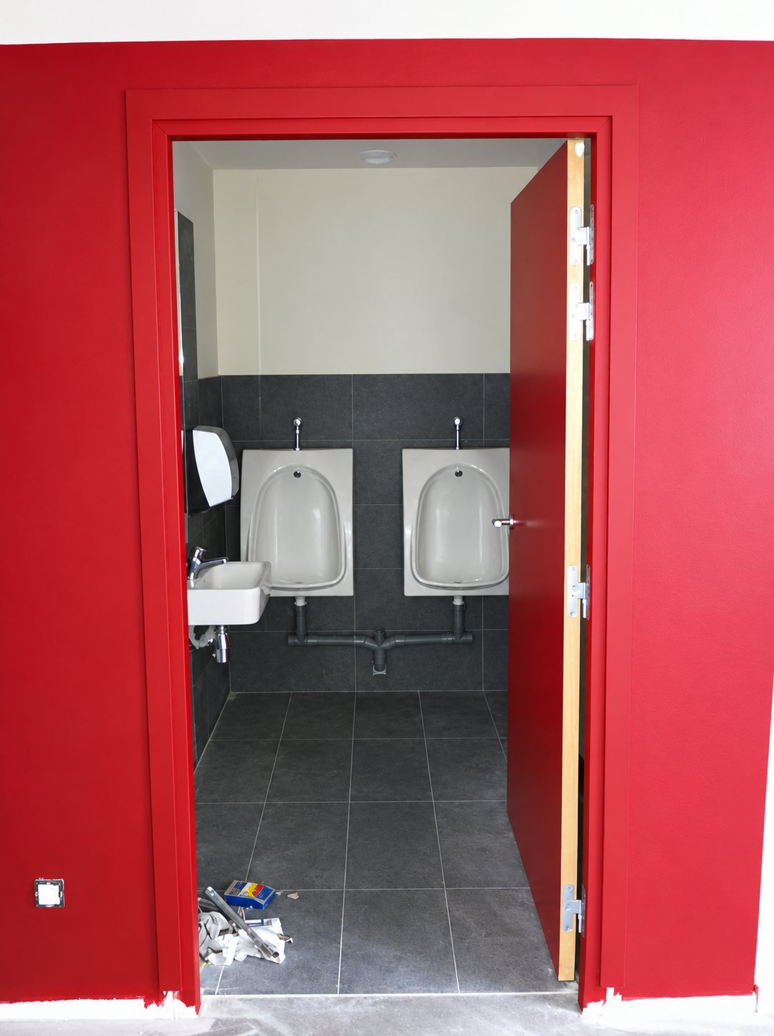 Porte ouverte dans un mur rouge donnant sur des toilettes carrelées de gris avec deux urinoirs et un lavabo, encore en travaux.