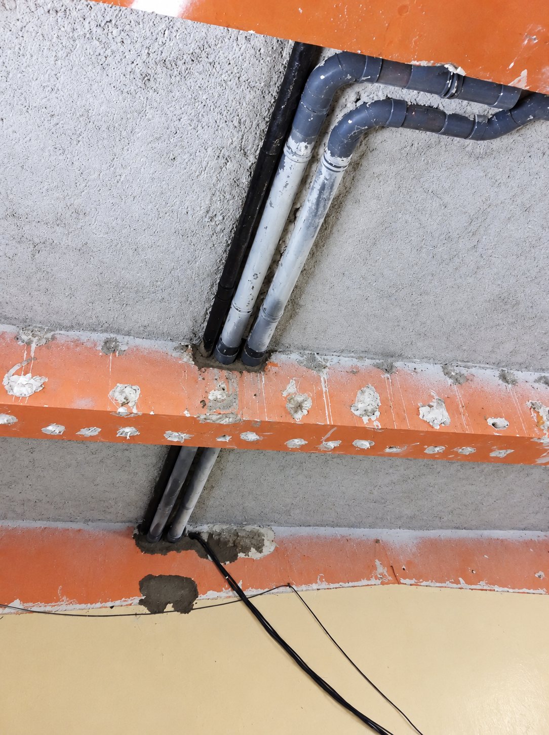 Vue en contre-plongée de deux tuyaux gris traversant un plafond en béton et des poutrelles en terre cuite, avec un câble noir qui pend.